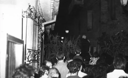 Przestrzeń miejska. Wąską uliczką pomiędzy budynkami przechodzi tłum ludzi. Widać tylko ich głowy. Na dalszym planie widać duże rośliny.