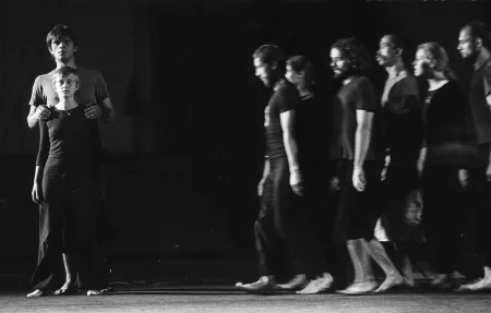 Po prawej stronie grupa aktorów i aktorek zwrócona do nas profilem w szeregu idzie w lewą stronę. Są ubrani w czarne spodenki i czarne podkoszulki. Są boso. Po lewej stronie stoi zwrócona twarzą w nasza stronę aktorka. Tuż za nią stoi zwrócony twarzą do nas wyższy od niej o głowę aktor obejmujący jej ramiona. Oboję są ubrani w czarne spodnie i ciemne podkoszulki. Są boso.