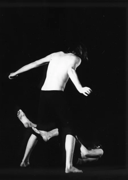 Po lewej stronie, tyłem do nas stoi aktor. Ma długie włosy, czarne spodenki i nie ma koszulki. Drugi aktor oplata jego nogi swoimi nogami i leży na podłodze.