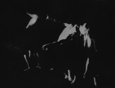 Niewyraźne, rozmazane zdjęcie. Troje aktorów stoi jeden za drugim. Każdy aktor trzyma za głowę aktora stojącego obok niego. Aktor stojący w głębi sceny jest zwrócony do nas plecami. Aktor stojący pośrodku wygina się, jakby chciał isć w głąb sceny. Za głowę trzyma go aktorka zwrócona do nas przodem.