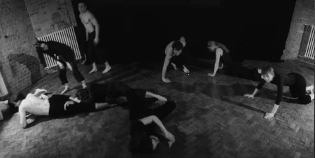 Grupa aktorów i aktorek tarza się po podłodze. Niektórzy są ubrani w czarne spodenki i czarne podkoszulki. Niektórzy nie mają podkoszulek. Są boso. Znajdują się na podłodze z paneli. W tle widać drzwi i kaloryfery.