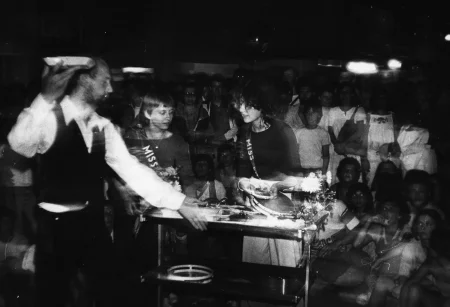 Aktor stoi przed dużą grupą widzów. Pokazuje stolik, na którym leżą przedmioty wyglądające na kwiaty i tamburyna.