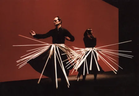 Na tle czerwonej ściany plecami do siebie stoją aktor i aktorka. Oboje ubrani są w czarne spódnice i czarna koszule. Na ubraniach mają zawieszone białe unoszące się patyki.