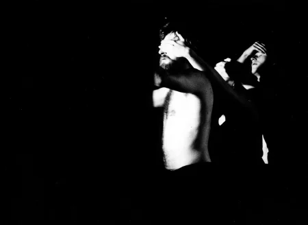 Aktor stoi bez koszuli i ma ręce wyciągnięte przed siebie. Jego oczy są zasłonięte przez dłoń osoby stojącej za nim. Po prawej stronie stoi aktor, który ma zamknięte oczy i trzyma rękę na czole.