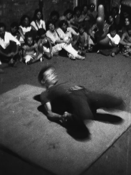 Niewyraźne zdjęcie. Na macie w gimnastycznej pozie leży aktor. Ma ugięte nogi, podpiera sie na łokciach. Na drugim planie siedzi grupa widzów.