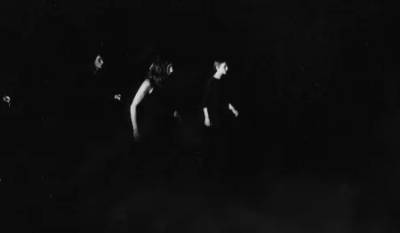 Niewyraźne zdjęcie. Z cienia wyłania się grupa aktorek stojących profilem w stronę widza. Aktorki ubrane są na czarno.