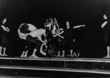 Na środku pustej sceny grupa aktorów wykonuje akrobacje. Są w bliskim kontakcie cielesnym. Po lewej i prawej stronie stoją zwróceni do nas profilem bracia bliźniacy ubrani w czarne podkoszulki i czarne spodenki. Są boso. Patrzą na siebie. Na środku, tuż przy grupie aktorów stoi aktorka wskazująca palcem na jednego z braci stojącego po naszej prawej stronie. Jest ubrana tak samo jak dwaj bracia.