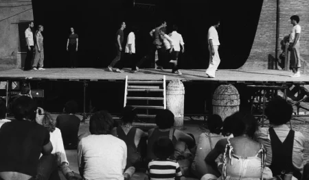 Scena plenerowa. Na scenę prowadzą drewniane schody. Z tyłu sceny ściana z czarnej płachty. Pod sceną siedzi grupa widzów. Po bokach sceny grupa aktorów i aktorek. Wyglądają, jakby przechodzili z jednego boku sceny na drugi. Wyróżnia się jedna aktorka stojąca z tyłu sceny po lewej stronie. Aktorka zwrócona jest twarzą w stronę widza. Ubrana jest w czarne spodnie i czarną podkoszulkę.