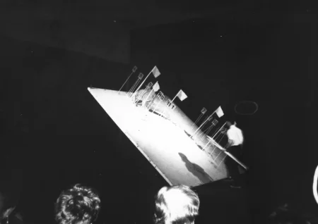 Na pierwszym planie widać głowy publiczności odwróconej plecami do widza. Przed publicznością stoi przechylony biały kwadrat z wbitymi białymi flagami i cyferkami. Obok kwadratu stoi odwrocony plecami do widza aktor. W ręce trzyma flagi i cyferki do wbijania w kwadrat.