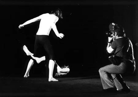 Po lewej stronie, tyłem do nas stoi aktor. Ma długie włosy, czarne spodenki i nie ma koszulki. Drugi aktor oplata jego nogi swoimi nogami i leży na podłodze. Nagrywa ich kamerzysta stojący po prawej stronie, tyłem do nas. Kamerzysta ubrany jest w podkoszulek i dżinsy.