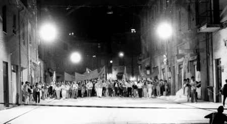 Przestrzeń miejska. Między budynkami stoi tłum ludzi zwróconych twarzą w stronę widza. Niektórzy trzymają w rękach białe flagi.