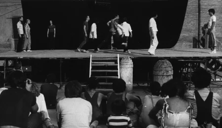 Scena plenerowa. Na scenę prowadzą drewniane schody. Z tyłu sceny ściana z czarnej płachty. Pod sceną siedzi grupa widzów. Po bokach sceny grupa aktorów i aktorek. Wyglądają, jakby przechodzili z jednego boku sceny na drugi. Wyróżnia się jedna aktorka stojąca z tyłu sceny po lewej stronie. Aktorka zwrócona jest twarzą w stronę widza. Ubrana jest w czarne spodnie i czarną podkoszulkę.