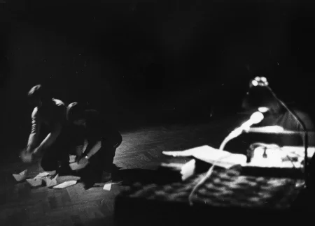 Po lewej stronie na ugiętych nogach stoją aktor i aktorka. Składają statki z papieru i ustawiają je na podłodze. Z prawej strony stoi biurko oświetlone stojącą lampką.