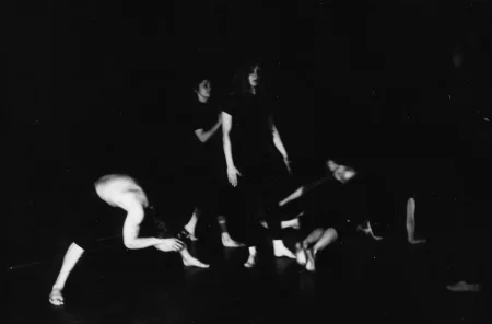 Niewyraźne, rozmazane zdjęcie. Po lewej stoi aktor. Jest ubrany w czarne spodenki, ma nagi tors. Jest boso. Schyla się ku podłodze, jakby się rozciągał. Po prawej stronie stoi i siedzi grupa aktorek. Są ubrane na czarno, są boso.