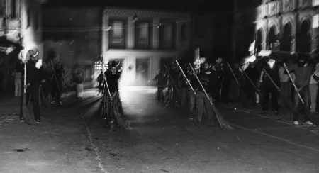 Niewyraźne zdjęcie. Przestrzeń miejska. Między budynkami w rzędach stoi grupa aktorów i aktorke. Trzymają w rękach miotły, którymi zamiatają ulicę.