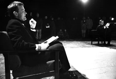 Na pierwszym planie na krześle siedzi aktor zwrócony profilem w stronę widza. Jest ubrany elegancko i coś mówi. Na kolanach trzyma książkę. Po prawej stronie na krześle siedzi aktor zwrócony przodem w stronę widza. Ustawia mikrofon. Za jego plecami stoi publiczność zwrócona przodem w stronę widza. Pomiędzy aktorami leży biała płachta.