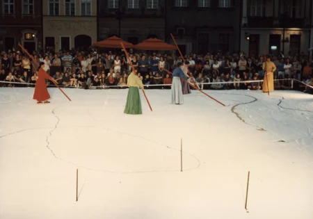 Duża biała scena ustawiona w przestrzeni miejskiej. Scena wygląda, jakby był na niej śnieg. Na scenie stoi czterosoobowa grupa aktorów i aktorek ubranych w kolorowe suknie w stylu azjatyckim. Trzymają w rękach długie czerwone kije i rysują wzory na scenie. W tle pod budynkami stoi publiczność zwrócona twarzą w stronę widza.