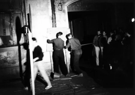 Przestrzeń miejska. Pod ścianą budynku dwóch aktorów zwróconych plecami w stronę widza ustawia tudny do określenia przedmiot. Po lewej stronie idzie przechodzień. Po prawej stronie stoi grupa widzów patrząca na aktorów.