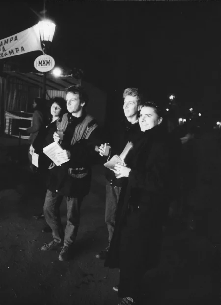 Dwóch aktorów i dwie aktorki. Uśmiechają się. Ubrani są w ciepłe ubranie, takie jak spodnie jeansowe i długie kurtki. Za nimi stoi latarnia i pusty stragan.