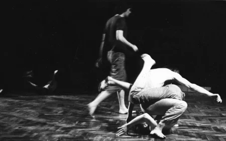 Dwóch splecionych ze sobą aktorów wykonuje akrobatyczną pozę siedząc na podłodze. Wyglądają, jakby byli jednym ciałem. Za nimi wzdłuż sceny przechodzi aktor. Ubrany jest w ciemne spodenki i ciemną podkoszulkę. Jest boso.