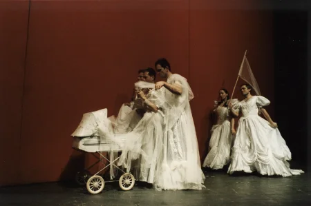 Na tle czerwonej ściane przez scenę przechodzi czterech aktorów i jedna aktorka. Wszyscy ubrani są w białe suknie ślubne. Trzech aktorów idzie z przodu. Pchają biały wózek dziecięcy. Za nimi idzie aktor i aktorka. Aktor trzyma w ręce białą flagę na kiju.