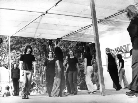 Grupa aktorów i aktorek na zadaszonej, plenerowej scenie. Na prawej ścianie sceny widać zasłonięty przez aktorki napis, prawdopodobnie "avant garde". Aktorzy i aktorki chodzą po scenie w kołko. Są ubrani w jeansy i czarne podkoszulki. W tle widać las, namioty i widownię.