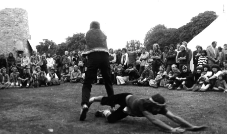Plener, ruiny jakiegoś budynku. W centru dwóch aktorów. Jeden aktor stoi na palcach. Ubrany jest w ciemne spodnie i kamizelkę. Pod jego nogami leży aktor. Wygląda, jakby upadał na ziemię. Ubrany jest w ciemne spodnie i podkoszulkę. W tle widać grupę widzów.