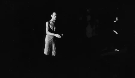 Niewyraźne zdjęcie. Z cienia wyłania się aktor stojący profilem w stronę widza. Ubrany jest w jasne spodenki, jego tors jest nagi. Po prawej stronie stoi słabo widoczna aktorka. Ubrana jest na czarno.