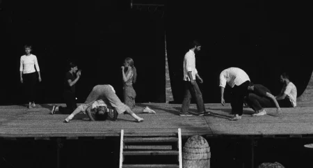 Scena plenerowa. Z tyłu sceny ściana z czarnej płachty. Po lewej stronie stoi aktorka ubrana w czarne spodnie i białą koszulkę z długim rękawem. Obok niej naprzeciwko siebie klęczą dwie aktorki. Pokazują sobie wnętrza dłoni. Pomiędzy nimi aktor lub aktorka wygląda, jakby robił/a fikołek. Z prawej strony czterech aktorów. Jeden z nich stoi, drugi jest pochylony, trzeci siada, czwarty siedzi z wyprostowanymi nogami.