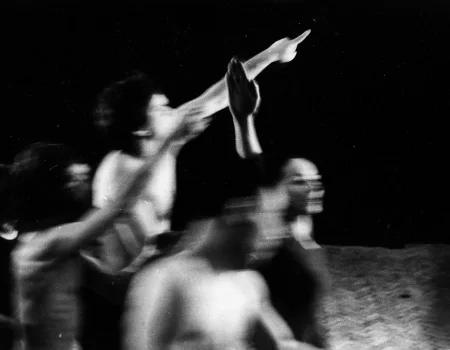 Niewyraźne, rozmazane zdjęcie. Grupa aktorów i aktorej w zwartej grupie. Każdy ma wyciągniętą w górę jedną rękę. Znajdują się na podłodze z paneli. W tle opuszczona czarna kurtyna.