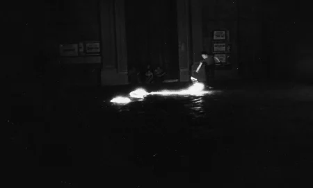 Niewyraźne zdjęcie. Przestrzeń miejska. Na ulicy stoi aktor. Wygląda, jakby coś podpalał. Za jego plecami widać budynek, pod którym siedzi troje widzów.