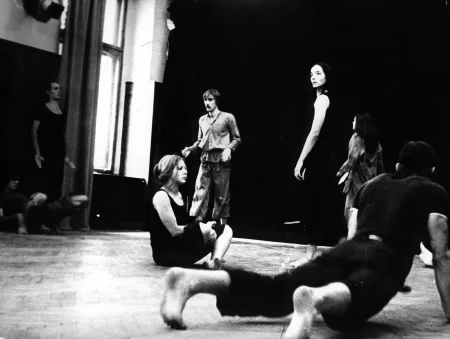 Grupa aktorów i aktorek w przestrzeni oświetlonej odsłoniętym oknem. Każdy wykonuje inną czynność, nie zwracają na siebie uwagi. Na pierwszym planie widać podeszwy stóp aktora leżącego na podłodze na brzuchu. W centrum jeden aktor i dwie aktorki. Jedna aktorka siedzi w siadzie skrzyżnym. Ubrana jest na czarno. Druga aktorka i aktor stoją patrząc w przestrzeń. Po lewej stronie pod ścianą obok okna stoi wyprostowany aktor.