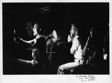 Przestrzeń miejska. Ulica wyglądająca jak tuż po zamieszkach. Pod zasłoniętymi witrynami sklepowymi leżą białe flagi. Ulicą idzie tłum ludzi.