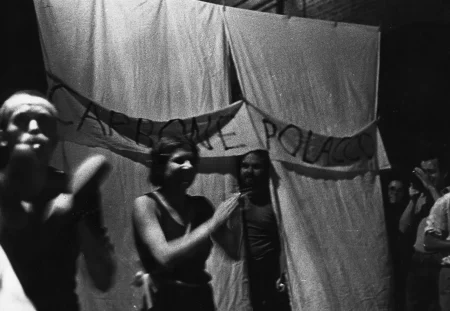 Grupa aktorów i aktorek. Trudno rozsztrzygnąć, kto jest kim. Za ich plecami wisi biała kurtyna, zza której wychodzi mężczyzna. Na kurtynie wisi biała szarfa z czarnym napisem "CARBONE POLACCO".