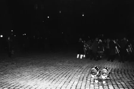 Niewyraźne zdjęcie. Przestrzeń miejska. Na brukowanej ulicy stoi grupa akorów i aktorek odwróconych do widza plecami. W rękach trzymają coś, co przypomina miotły. Po lewej stronie widać grupę widzów i przechodniów.