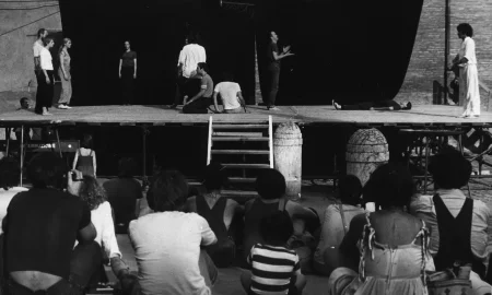 Scena plenerowa. Na pierwszym planie odwróceni plecami widzowie patrzą na scenę. Na scenę prowadzą drewniane schody. Z tyłu sceny ściana z czarnej płachty. Pośrodku, zza płachty wystaje latarnia. Na scenie stoją aktorzy i aktorki w różnych działaniach. Wyglądają, jakby mieli na sobie prywatne ubrania. Wspólnie tworzą choreograficzną kompozycję. Na dalszym planie, za sceną widać budynki.