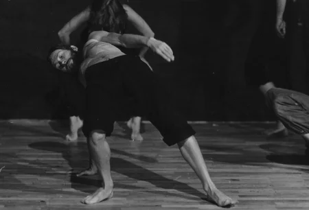 Aktor, ustawiony brzuchem do góry, podpiera się jedną ręką o podłogę. Ubrany jest w czarne spodenki, ma nagi tors. Jest boso. Patrzy w podłogę. Za jego plecami słabo widoczna aktorka stoi na palcach i zwisa głową w dół.