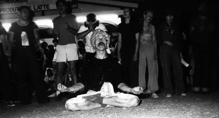 W centrum siedzi aktor w siadzie skrzyżnym. Wygląda, jakby medytował. Trzyma zgięte ręce po bokach, ma otwarte usta. Widać tylko jego białka oczu. Na głowie ma arafatkę. Za nim w szregu stoi grupa aktorów i aktorek. Wyglądają, jakby śpiewali.