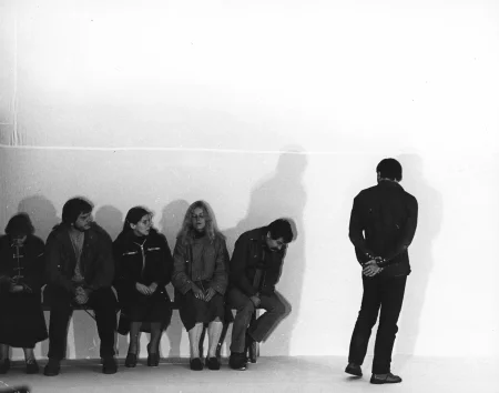 Biała sterylna przestrzeń. Pod ścianą na ławce siedzi grupa aktorów i aktorek zwróconych przodem w stronę widza. Obok ławki stoi aktor zwrócony w stronę widza plecami. Stojący aktor patrzy na aktorów na ławce.