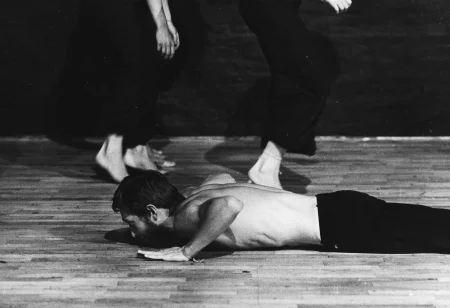 Aktor leży brzuchem na podłodze. Podpiera się rękami. Jego tors jest nagi. Za nim idą na palcach aktorki. Widoczne są tylko ich nogi. Ubrane są w czarne spodnie.