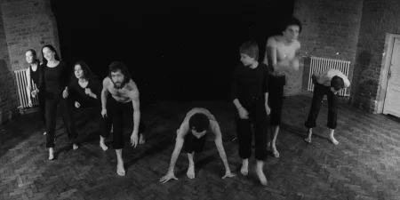 Aktorzy i aktorki zwróceni twarzą w stronę widza. Wszyscy są w ruchu. Wyglądają, jakby rozgrzewali się przed wysiłkiem. Aktorki ubrane są w czarne spodnie i czarne koszulki. Aktorzy są ubrani w czarne spodenki i mają nagie torsy. W tle widać czarną płachtę przykrywającą ceglaną ścianę. Po dwóch stronach płachty widać białe kaloryfery.