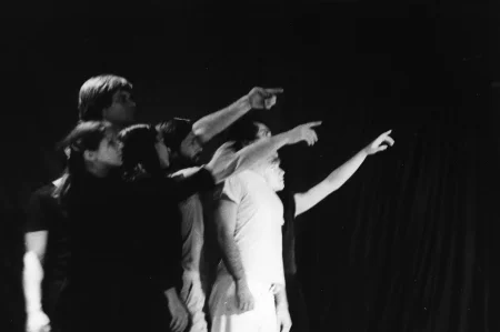 Grupa aktorów i aktorek stojących profilem w stronę widza. Wspólnie patrzą w jeden punkt i wskazują na niego palcem. Aktorzy i aktorki stoją na tle czarnej płachty.