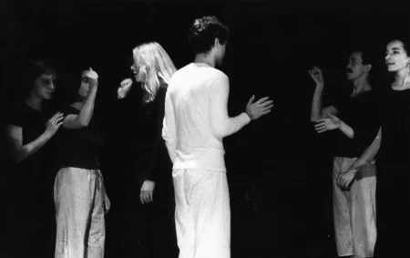 Aktorzy i aktorki patrzą na siebie i gestykulują w swoją stronę rękami. Wyróżnia się aktor stojący w centrum. Aktor stoi zwrócony plecami w stronę widza. Jest ubrany na biało.