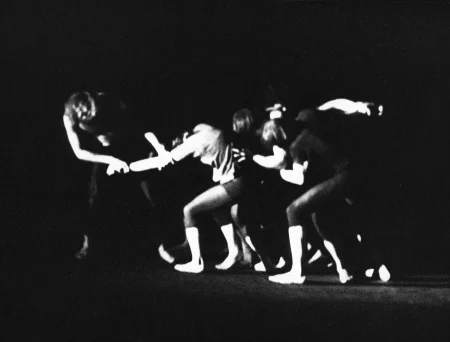 Niewyraźne, rozmazane zdjęcie. Po prawej stronie aktorzy garbią się i trzymają się wzajemnie. Po lewej stronie aktor trzyma za rękę aktora po prawej, jakby chciał przyciągnąć do siebie całą grupę.