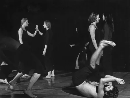 Grupa aktorów i aktorek. Na pierwszym planie dwóch aktorów wykonuje akrobatyczne ruchy na podłodze. Za nimi, w dwóch parach stoją cztery aktorki. W każdej parze aktorki są zwrócone do siebie twarzami. W oddali przechodzi przez scenę słabo widoczny aktor. Aktorzy ubrani są w czarne spodenki, mają nagie torsy. Aktorki ubrane są w czarne spodnie i czarne koszulki. Wszyscy są boso.