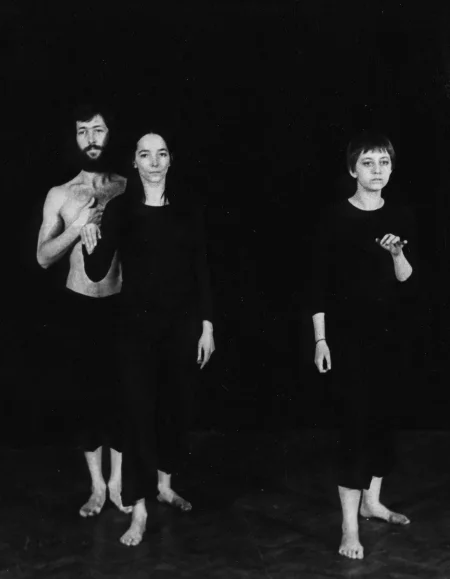 Jeden aktor i dwie aktorki. Stoją zwróceni twarzą w stronę widza, jakby pozowali do zdjęcia. Po lewej stronie aktor i aktorka stoją bardzo blisko siebie. Aktorka po prawej stronie stoi samotnie. Aktor ubrany jest w czarne spodenki i ma nagi tors. Aktorki ubrane są w czarne spodnie i czarne koszulki. Wszyscy są boso.