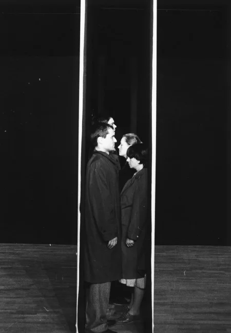 Grupa aktorów i aktorek stoi ściśnięta pomiędzy dwiema ścianami ustawionymi blisko na siebie na środku sceny. Stoją profilem w stronę widza i twarzą do siebie. Mają na sobie czarne płaszcze.