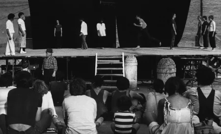 Scena plenerowa. Na pierwszym planie odwróceni plecami widzowie patrzą na scenę. Na scenę prowadzą drewniane schody. Z tyłu sceny ściana z czarnej płachty. Na scenie stoją aktorzy i aktorki w różnych działaniach. Wyglądają, jakby mieli na sobie prywatne ubrania. Wspólnie tworzą choreograficzną kompozycję. Aktorzy i aktorki po bokach sceny są statyczni. Aktorzy w centrum ruszają się dynamicznie. Na dalszym planie, za sceną widać budynki.