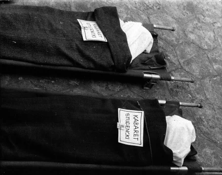 Dwie pary noszy wyglądających, jakby leżał na nich ktoś martwy przykryty kocem. Na noszach leżą tabliczki z napisem "KABARET STUDENCKI".