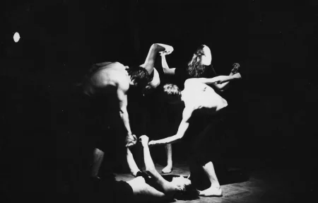 Aktor w czarnych spodenkach bez koszulki leży na podłodze. Trzyma się za ręcę z grupą aktorów, którzy też trzymają się za ręce. Mają ciała zwrócone w różne strony, razem tworzą okrąg. Dwóch aktorów w grupie ubranych jest w czarne spodenki, nie mają na sobie koszulek. Aktorka z grupy stoi zwrócona do nas tyłem. Ma spięte włosy.